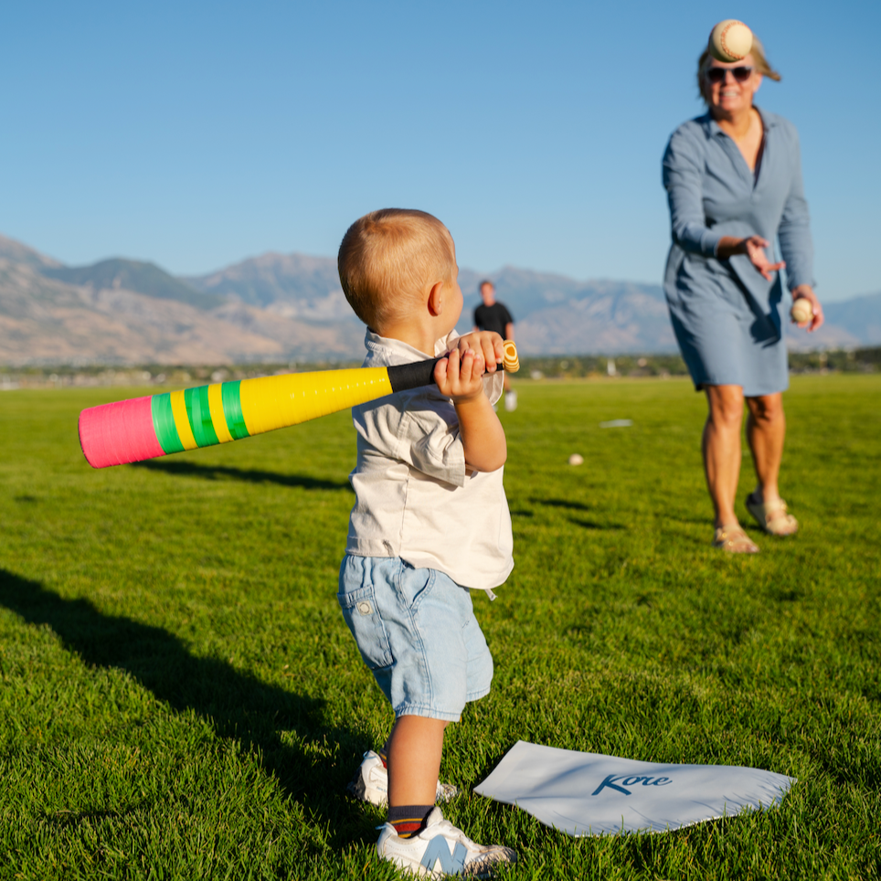 Child holding a pencil baseball bat on a grassy field with mountains in the background